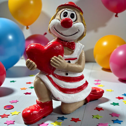 Clown-Figur mit roter Nase in rot-weißem Outfit, kniend dargestellt und ein rotes Herz haltend, auf einer Tischfläche mit Konfetti, im Hintergrund farbige Luftballons.