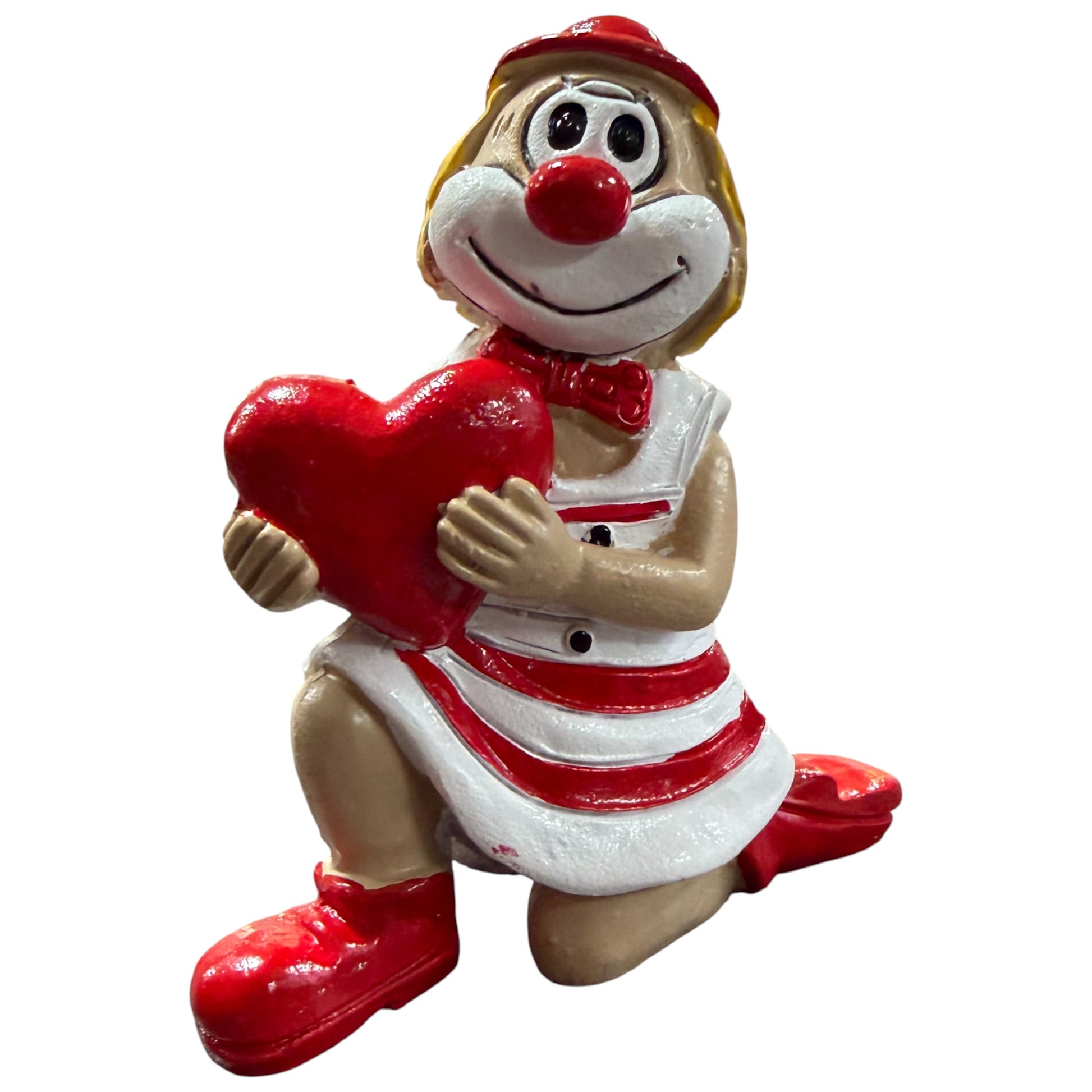 Clown-Figur mit roter Nase in rot-weißem Outfit, kniend dargestellt und ein rotes Herz vor dem Körper haltend, vor weißem Hintergrund.