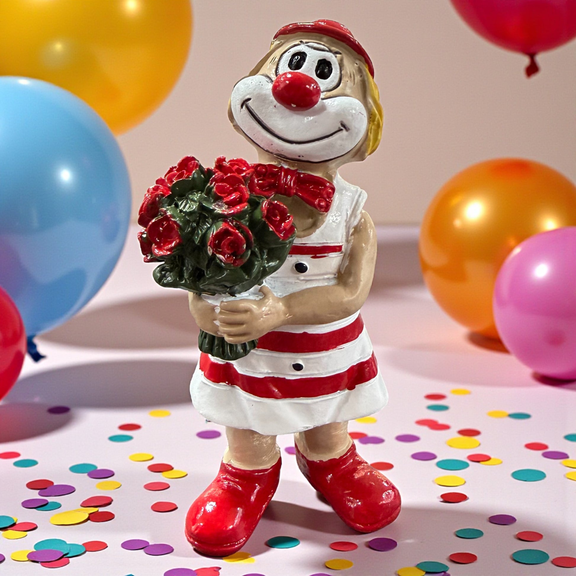 Clown-Figur mit roter Nase in rot-weißem Outfit, hält einen Blumenstrauß mit roten Rosen, stehend auf einer Tischfläche mit Konfetti, im Hintergrund farbige Luftballons.