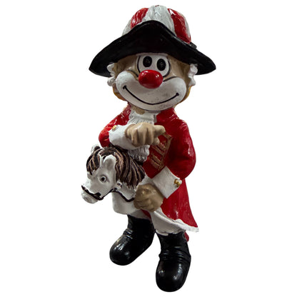 Clown-Figur mit roter Nase in roter Uniform und schwarzem Hut, hält ein kleines Pferd vor dem Körper, vor weißem Hintergrund.