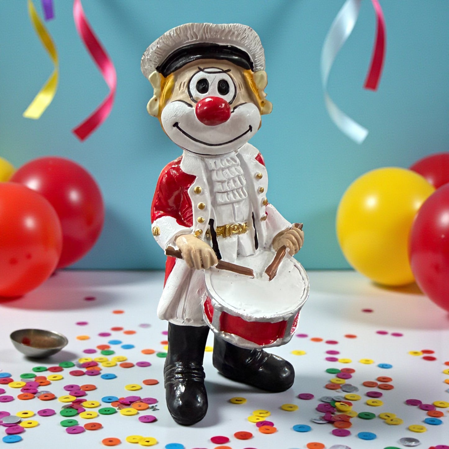Clown-Figur mit roter Nase in rot-weißer Uniform, hält eine Trommel und Trommelstöcke, stehend auf einer Tischfläche mit Konfetti, im Hintergrund Luftballons und bunte Bänder.