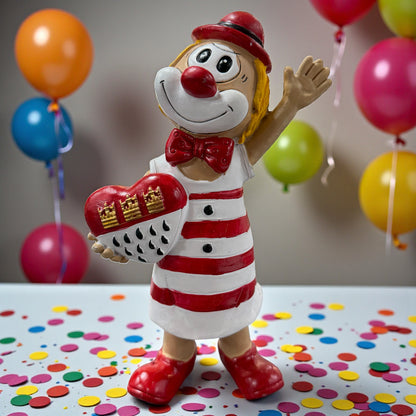 Große Clown-Dekofigur mit roter Nase in rot-weißem Outfit, ca. 20 cm groß, stehend dargestellt und ein Herz mit kölschem Stadtwappen haltend, auf einer Tischfläche mit Konfetti, im Hintergrund bunte Luftballons