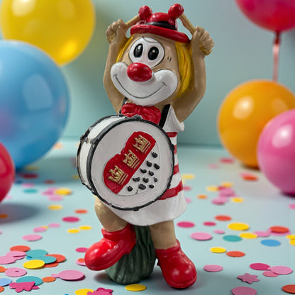 Große Clown-Dekofigur mit roter Nase in rot-weißem Outfit, ca. 20 cm groß, stehend dargestellt und eine Trommel mit kölschem Herzmotiv haltend, auf einer Tischfläche mit Konfetti, im Hintergrund bunte Luftballons.