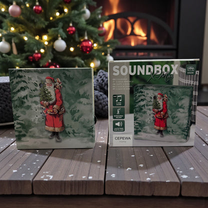 Eine weihnachtliche Soundbox mit Weihnachtsmann-Motiv steht neben ihrer Verpackung auf einem Holztisch vor Kamin und Weihnachtsbaum.
