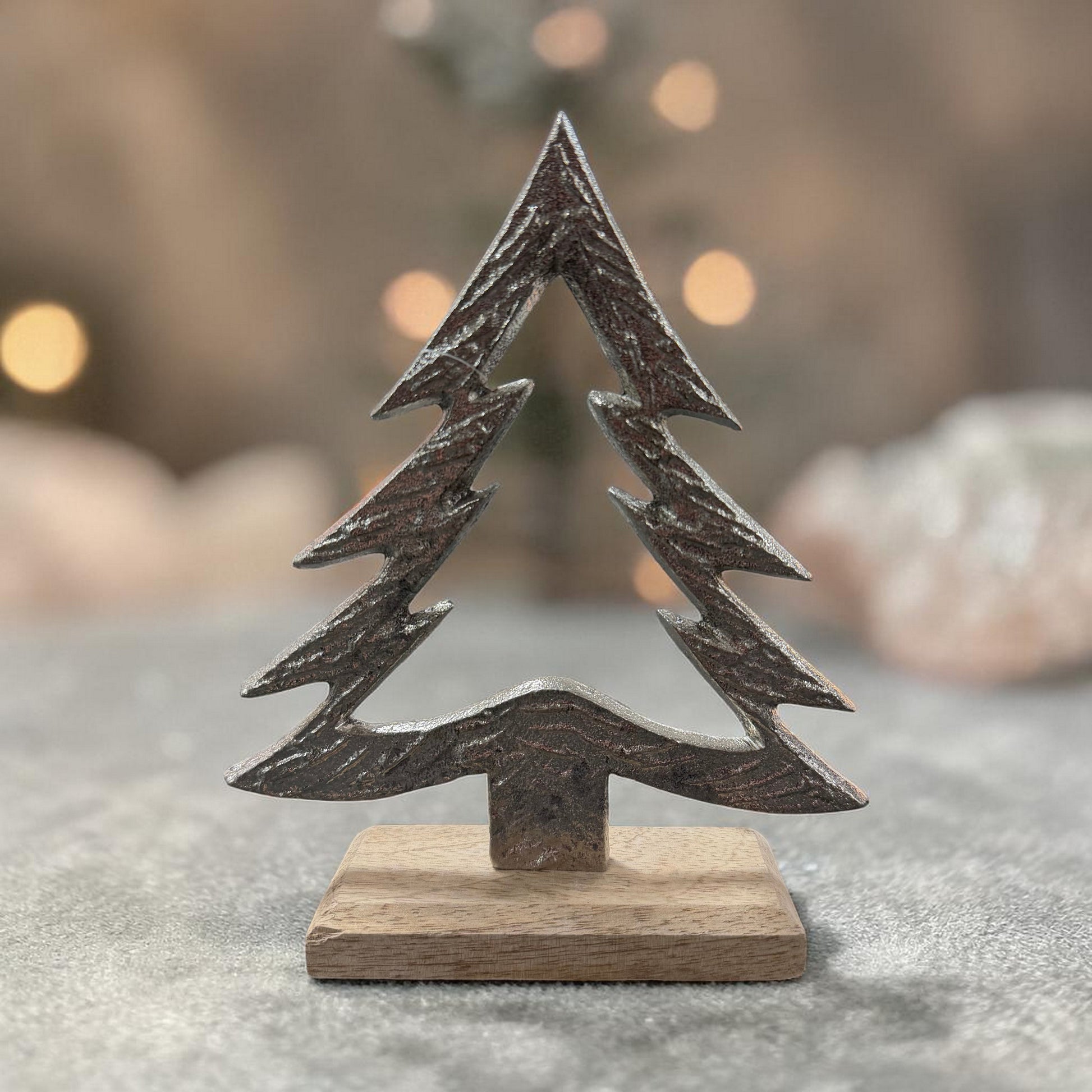 Ein stilisierter Tannenbaum aus dunklem Material steht auf einem rechteckigen Holzsockel vor einem unscharfen Hintergrund mit Lichtpunkten.