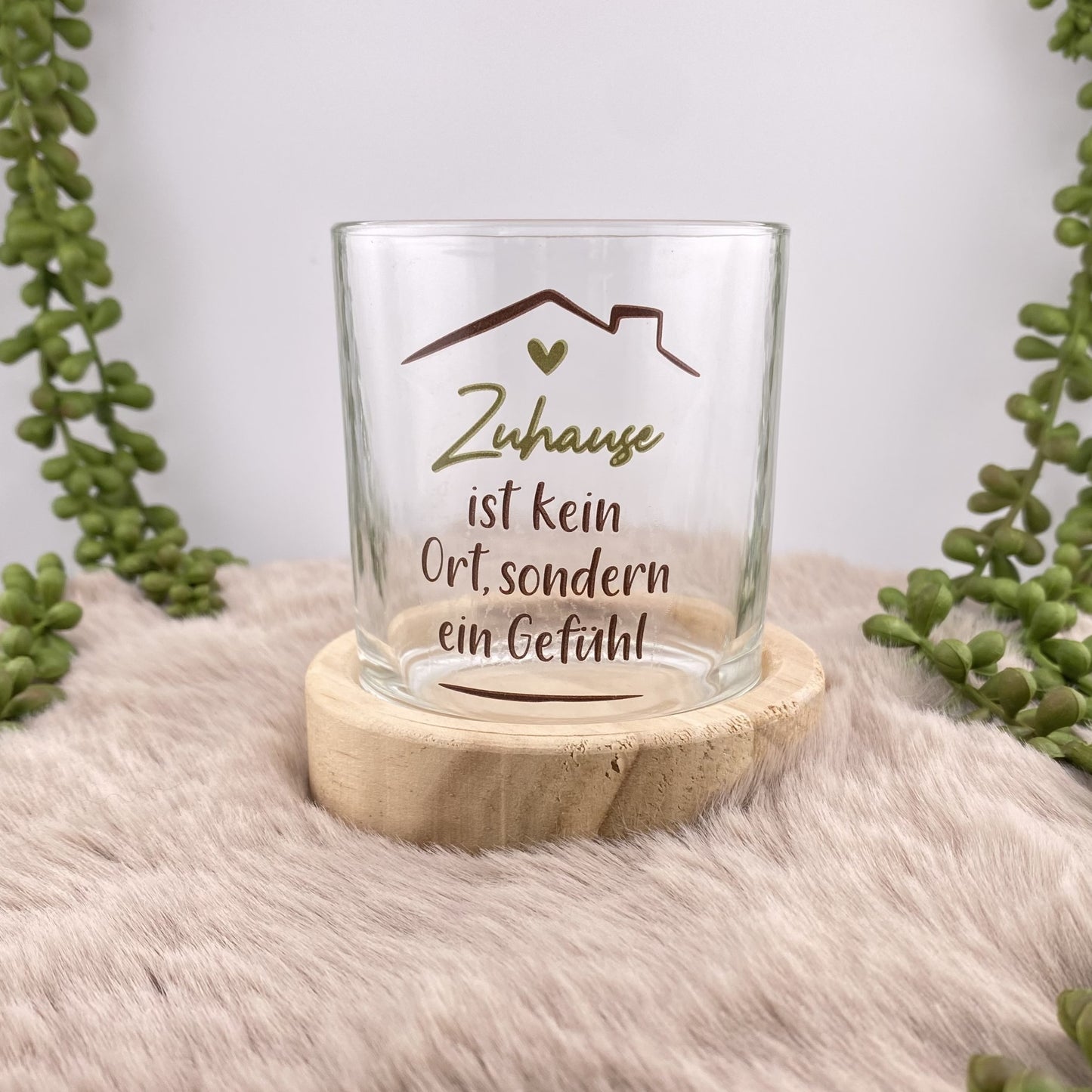 Ein Teelichthalter mit rundem Holzsockel und Glas mit olivem Schriftzug "Zuhause ist da, wo das Herz sich wohlfühlt" ist im Ambiente platziert