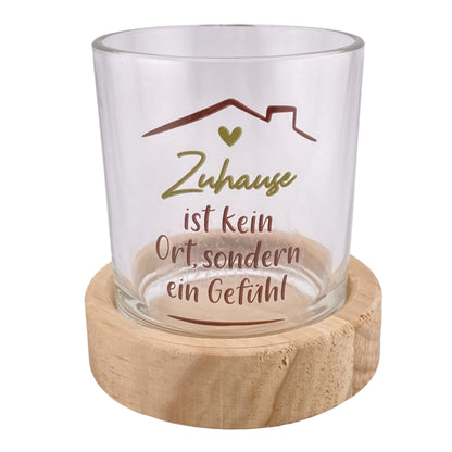 Ein Teelichthalter mit rundem Holzsockel und Glas mit olivem Schriftzug "Zuhause ist da, wo das Herz sich wohlfühlt" ist vor weißem Hintergrund platziert