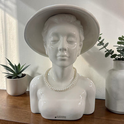 Eine weiße Keramik-Büste einer Frau mit großem Hut und Perlenkette steht auf einem Holztisch neben dekorativen Pflanzen und einer Vase vor einer hellen Wand.