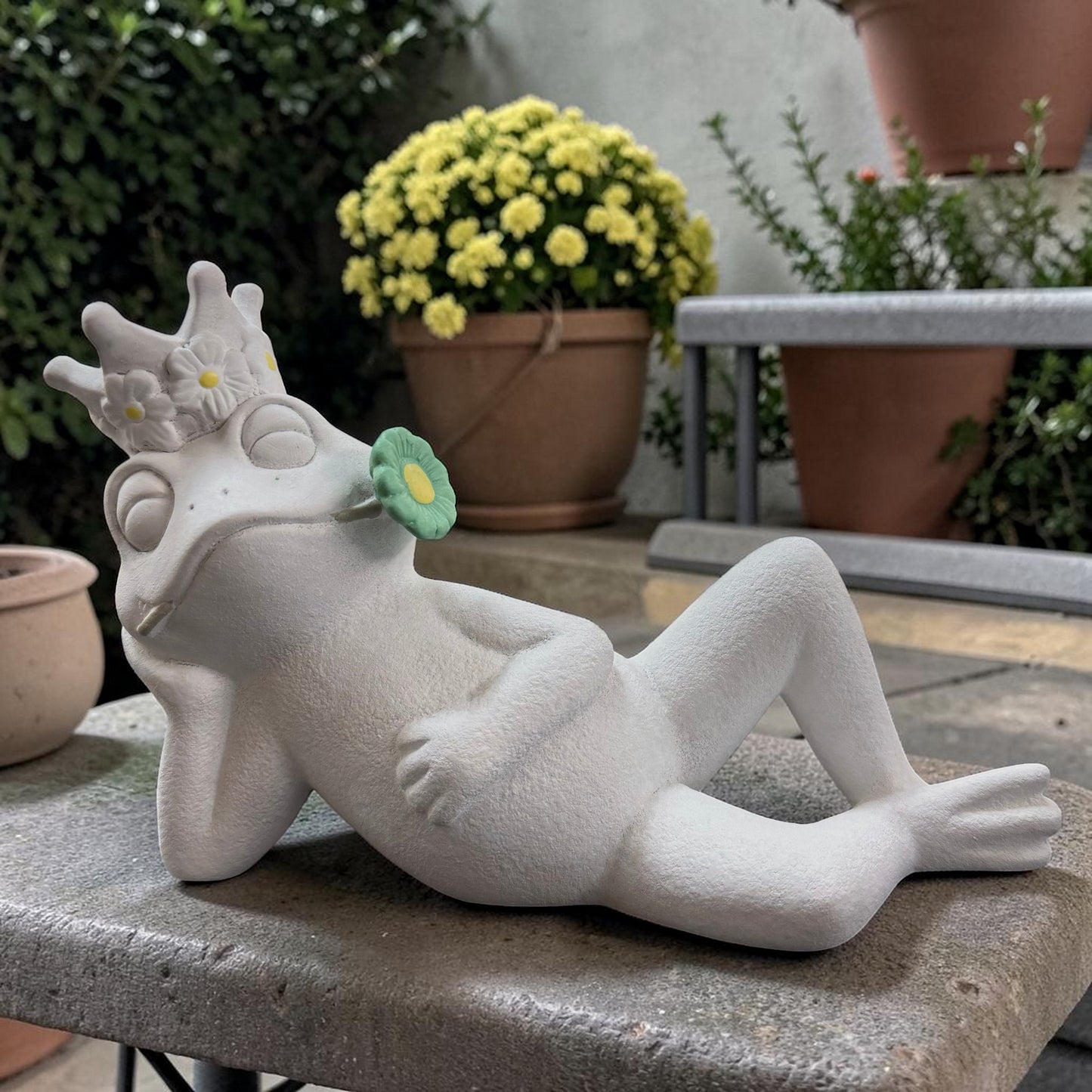 Großer weißer Deko-Frosch in liegender Haltung mit geschlossenen Augen und grüner Blüte im Mund.