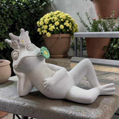 Großer weißer Deko-Frosch in liegender Haltung mit geschlossenen Augen und grüner Blüte im Mund.