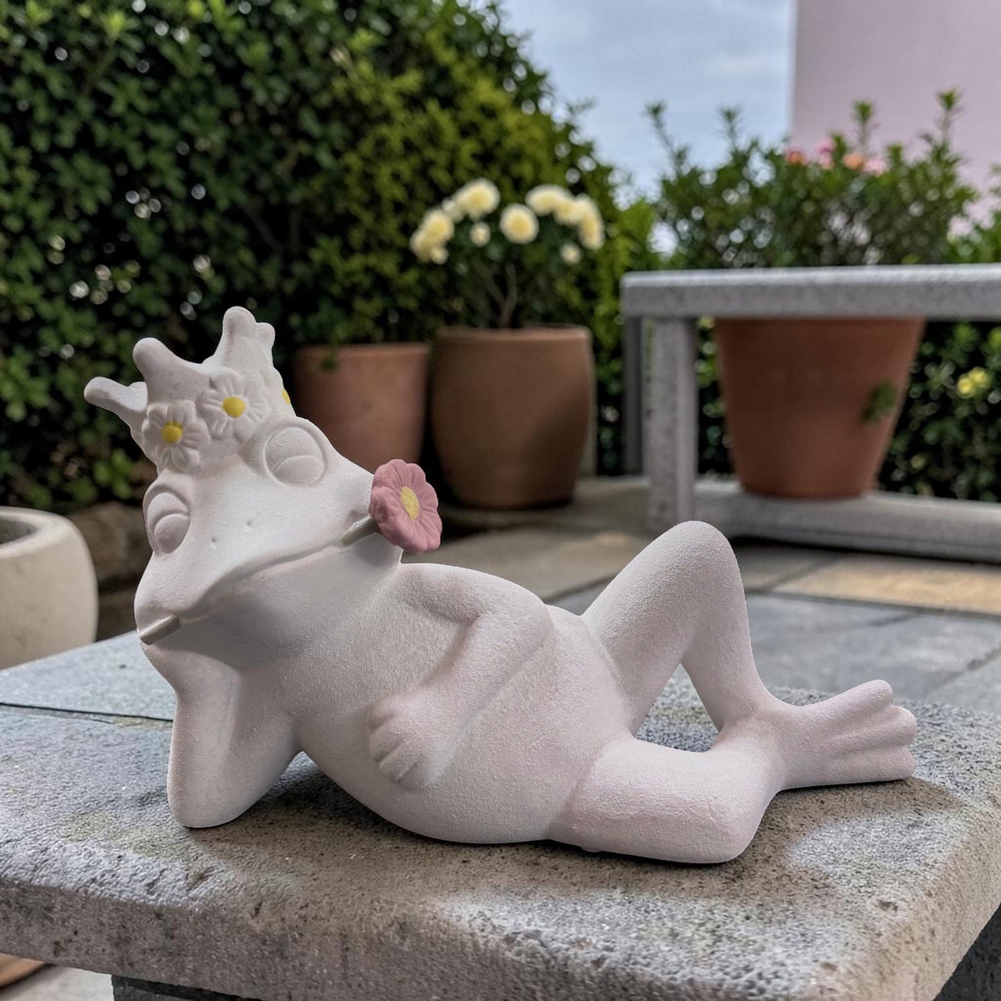 Weißer Deko-Frosch in liegender Haltung mit geschlossenen Augen und rosa Blüte im Mund.
