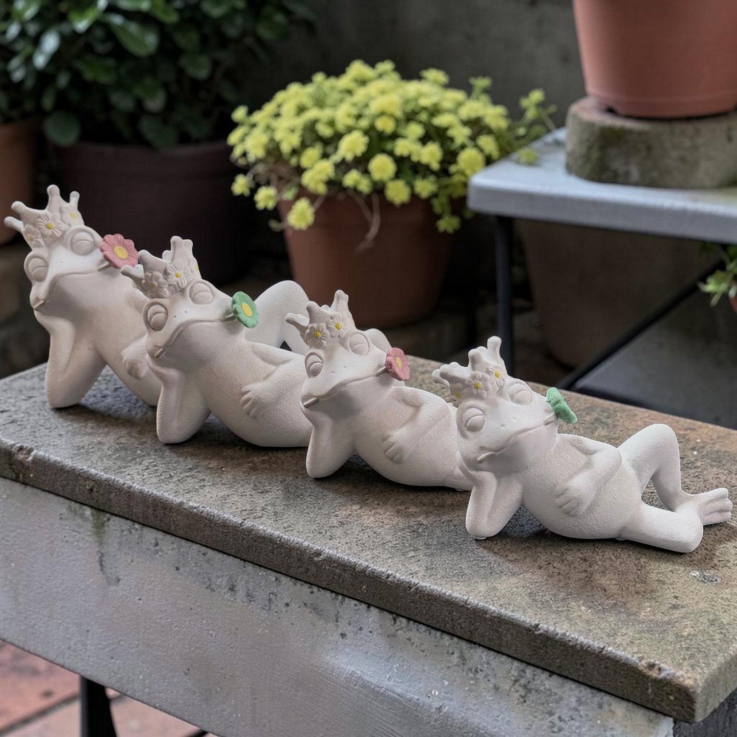 Vier liegende Froschfiguren in verschiedenen Größen mit Blumenkrone und farbigen Blüten im Ambiente