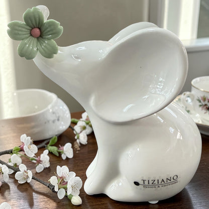 Ein weißer Keramik-Elefant mit grüner Blüte am Ohr ist auf einer Holzfläche mit Blütenzweigen und einer Tasse im Hintergrund arrangiert.