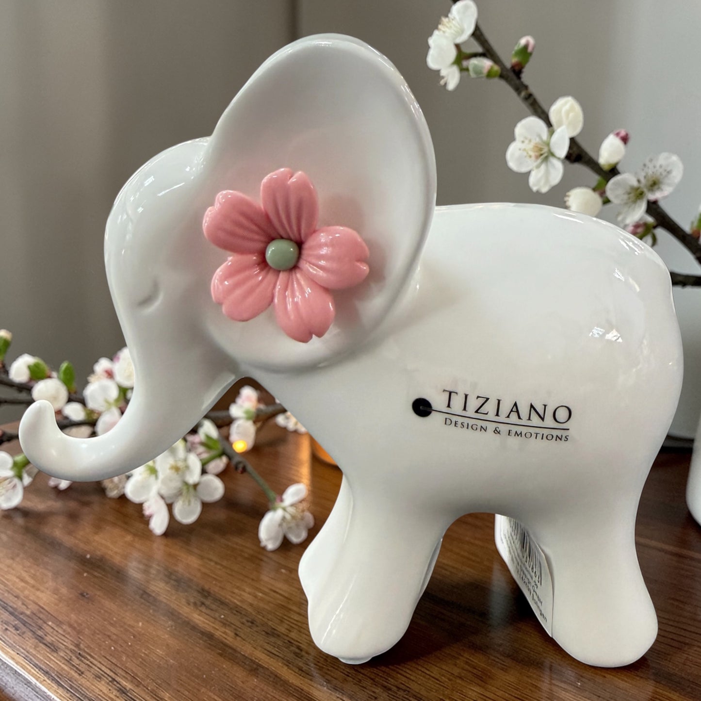 Ein weißer Keramik-Elefant mit rosa Blüte am Ohr steht auf einer Holzfläche neben blühenden Zweigen.