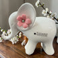 Ein weißer Keramik-Elefant mit rosa Blüte am Ohr steht auf einer Holzfläche neben blühenden Zweigen.