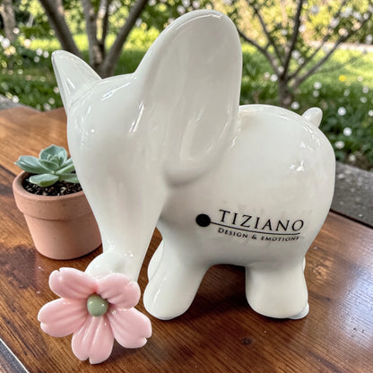 Ein weißer Keramik-Elefant mit rosa Blüte wird seitlich auf einer Holzoberfläche vor einem natürlichen Hintergrund gezeigt.