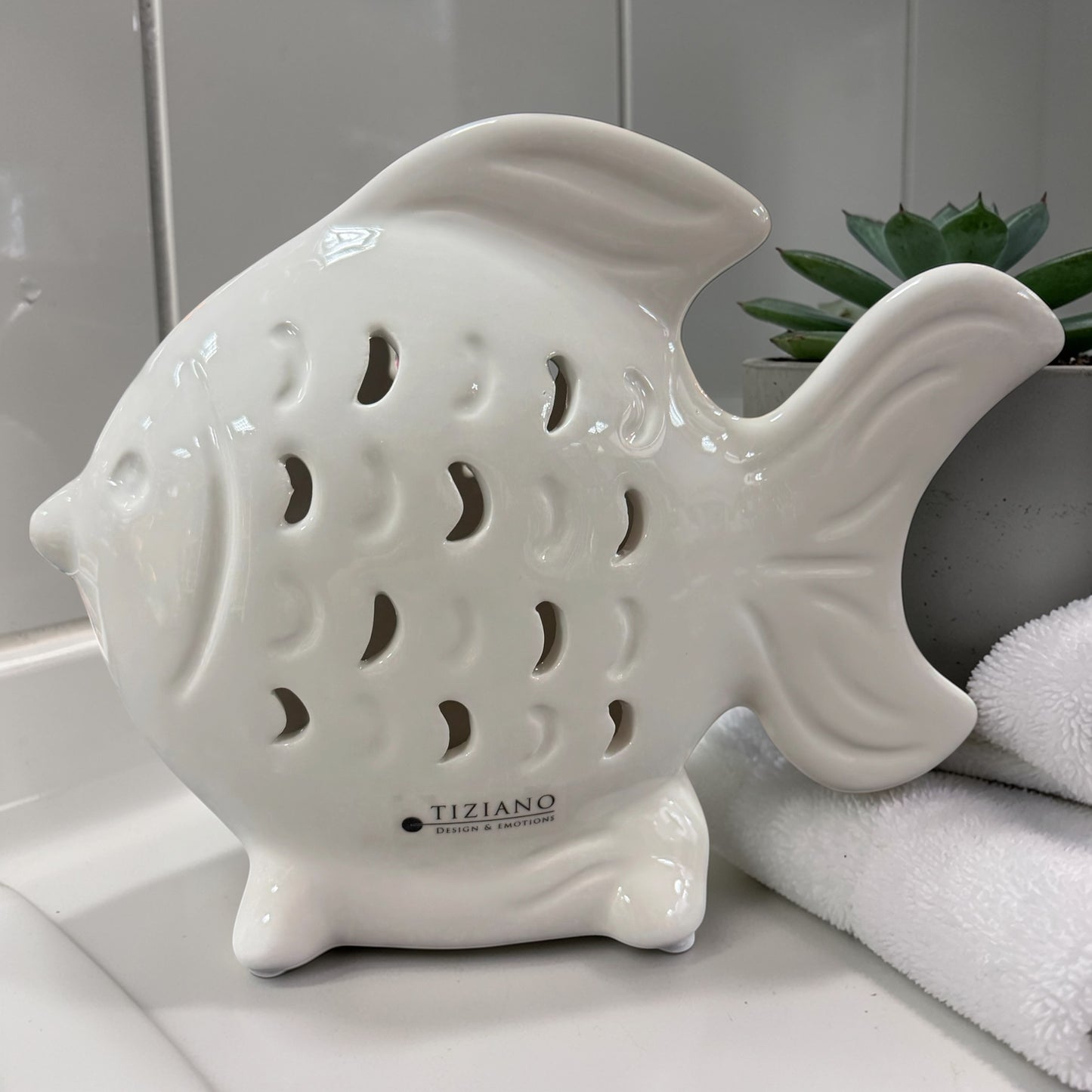 Ein weißer Keramik-Fisch mit dekorativen Ausstanzungen steht auf einer Badezimmerablage neben einem Handtuch und einer kleinen Pflanze.