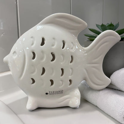 Ein weißer Keramik-Fisch mit dekorativen Ausstanzungen steht auf einer Badezimmerablage neben einem Handtuch und einer kleinen Pflanze.