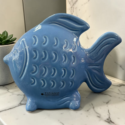 Ein großer blauer Keramik-Fisch wird als dekoratives Objekt auf einer Marmoroberfläche mit Pflanze im Hintergrund präsentiert.