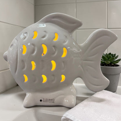 Ein weißer Keramik-Fisch mit integrierter LED-Beleuchtung leuchtet warm durch die ausgestanzten Muster und steht auf einer Badezimmerablage.