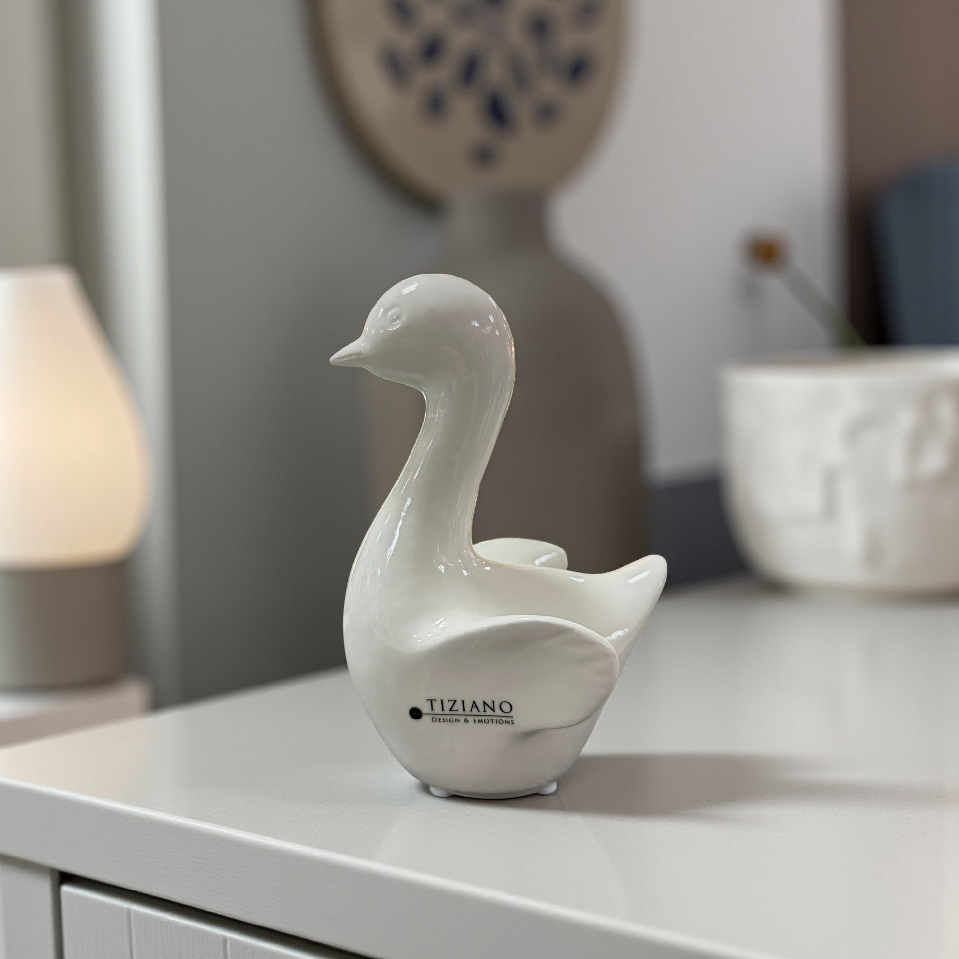 Weiße Keramikfigur in Gansform steht einzeln auf einem Möbelstück vor dekorativen Wohnaccessoires.