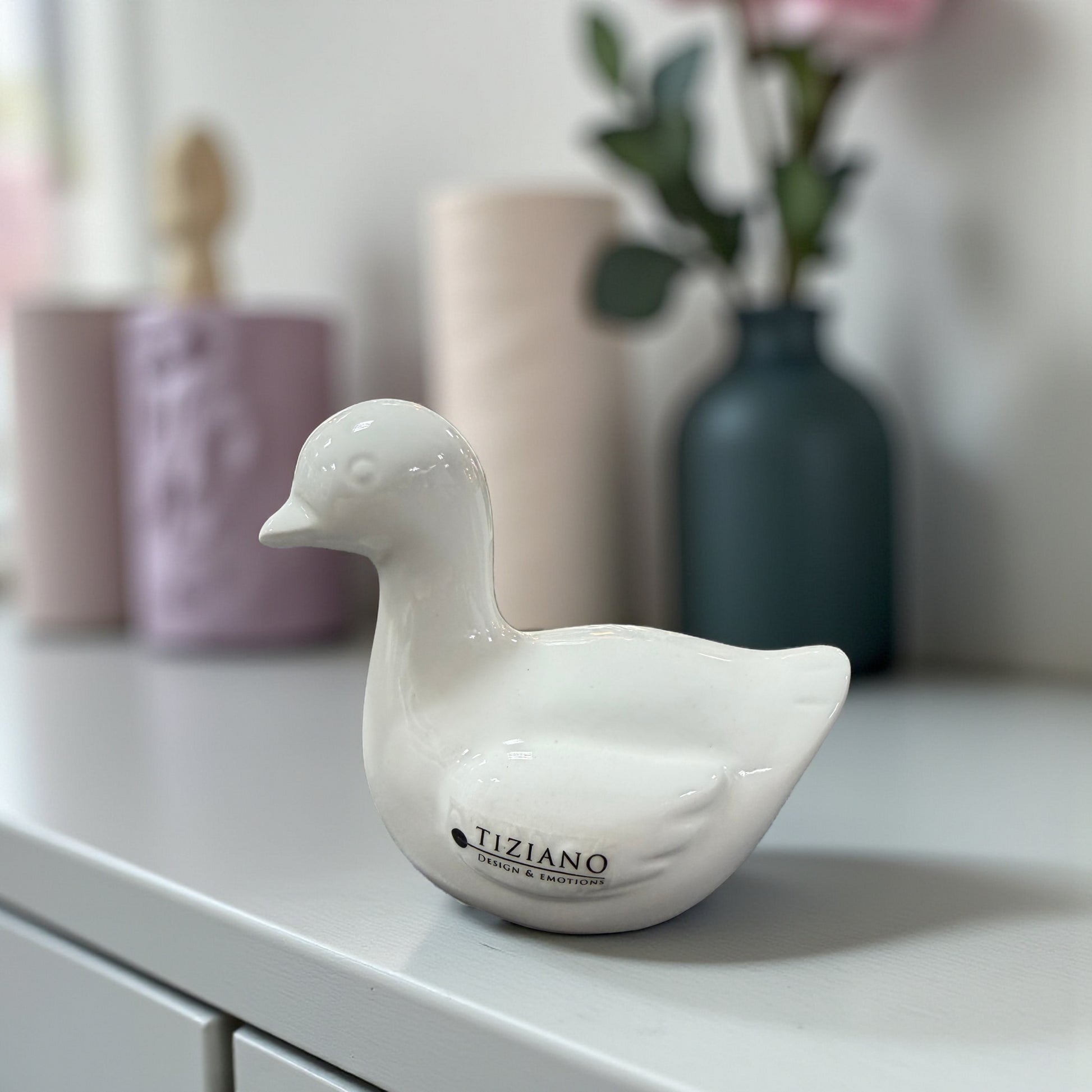 Kleine weiße Keramikfigur in Gansform steht auf einem Sideboard vor unscharfem Wohnhintergrund.