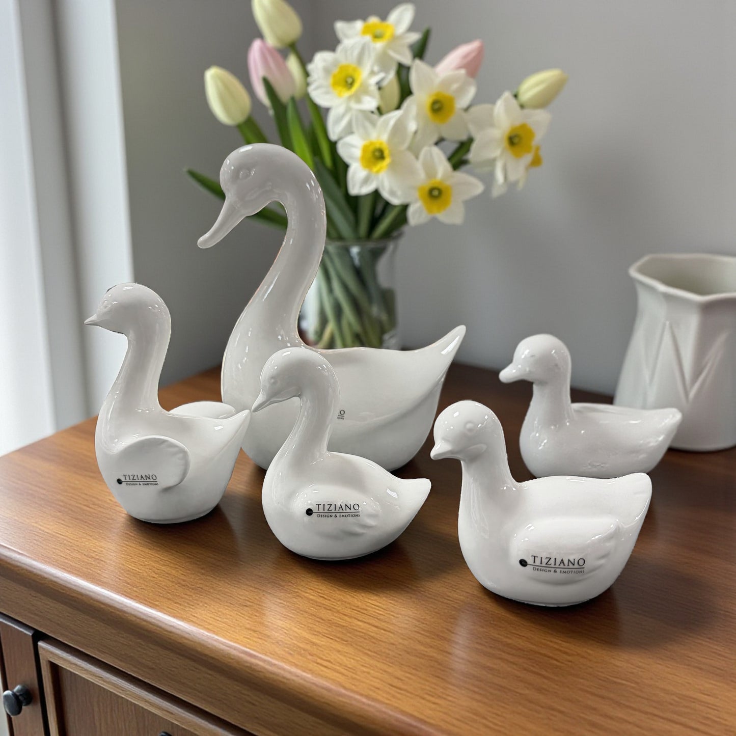 Mehrere weiße Keramik-Gänsefiguren in unterschiedlichen Größen stehen auf einem Holzsideboard vor Blumen.