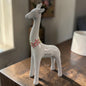 Eine weiße Keramik-Giraffe mit rosa Blumenkranz steht auf einem Holztisch vor einem unscharfen Wohnraumhintergrund mit Blumen und Kissen.