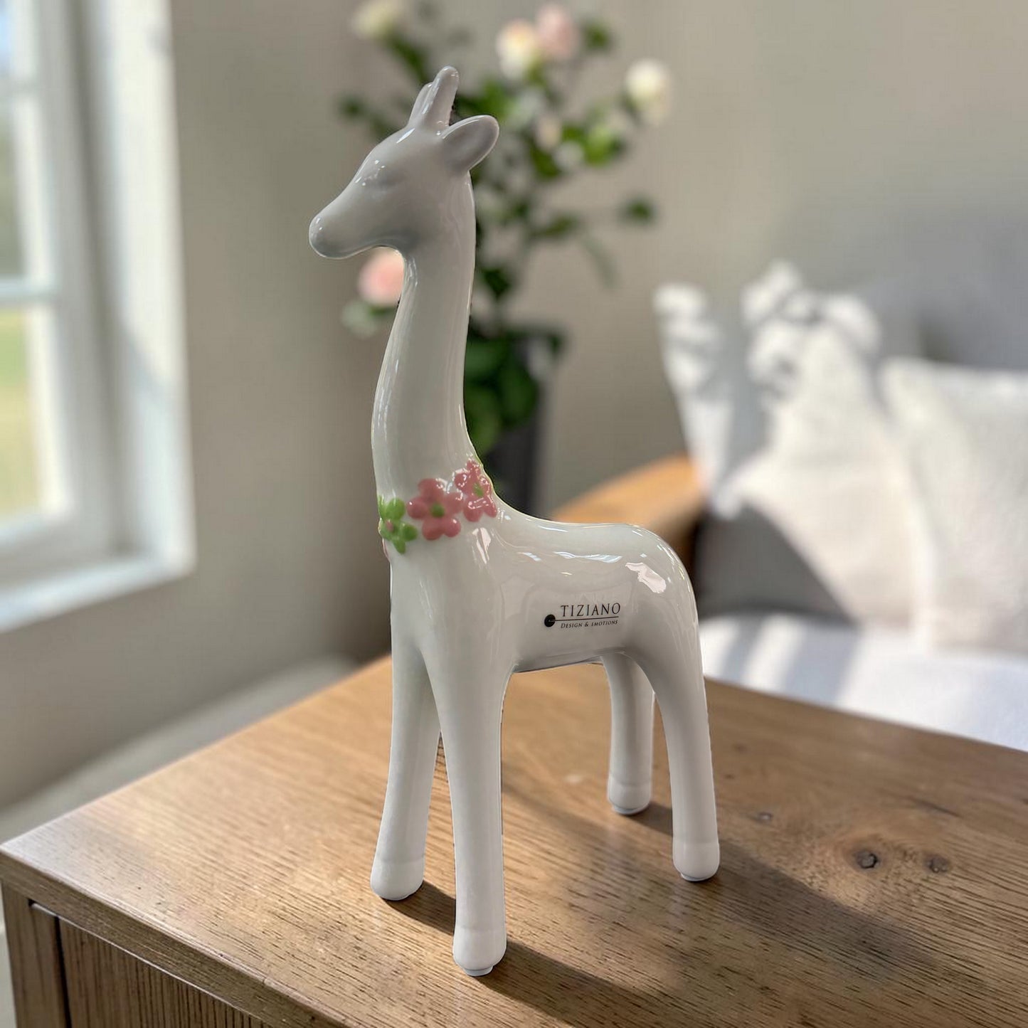 Eine weiße Keramik-Giraffe mit glänzender Oberfläche und rosa-grünem Blumenkranz steht auf einer dunklen Holzoberfläche vor einem hellen Wohnraumhintergrund.