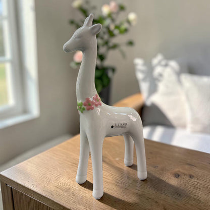 Eine weiße Keramik-Giraffe mit glänzender Oberfläche und rosa-grünem Blumenkranz steht auf einer dunklen Holzoberfläche vor einem hellen Wohnraumhintergrund.