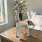 Eine weiße Keramik-Giraffe mit glänzender Oberfläche und rosa-grünem Blumenkranz steht auf einer dunklen Holzoberfläche vor einem hellen Wohnraumhintergrund.