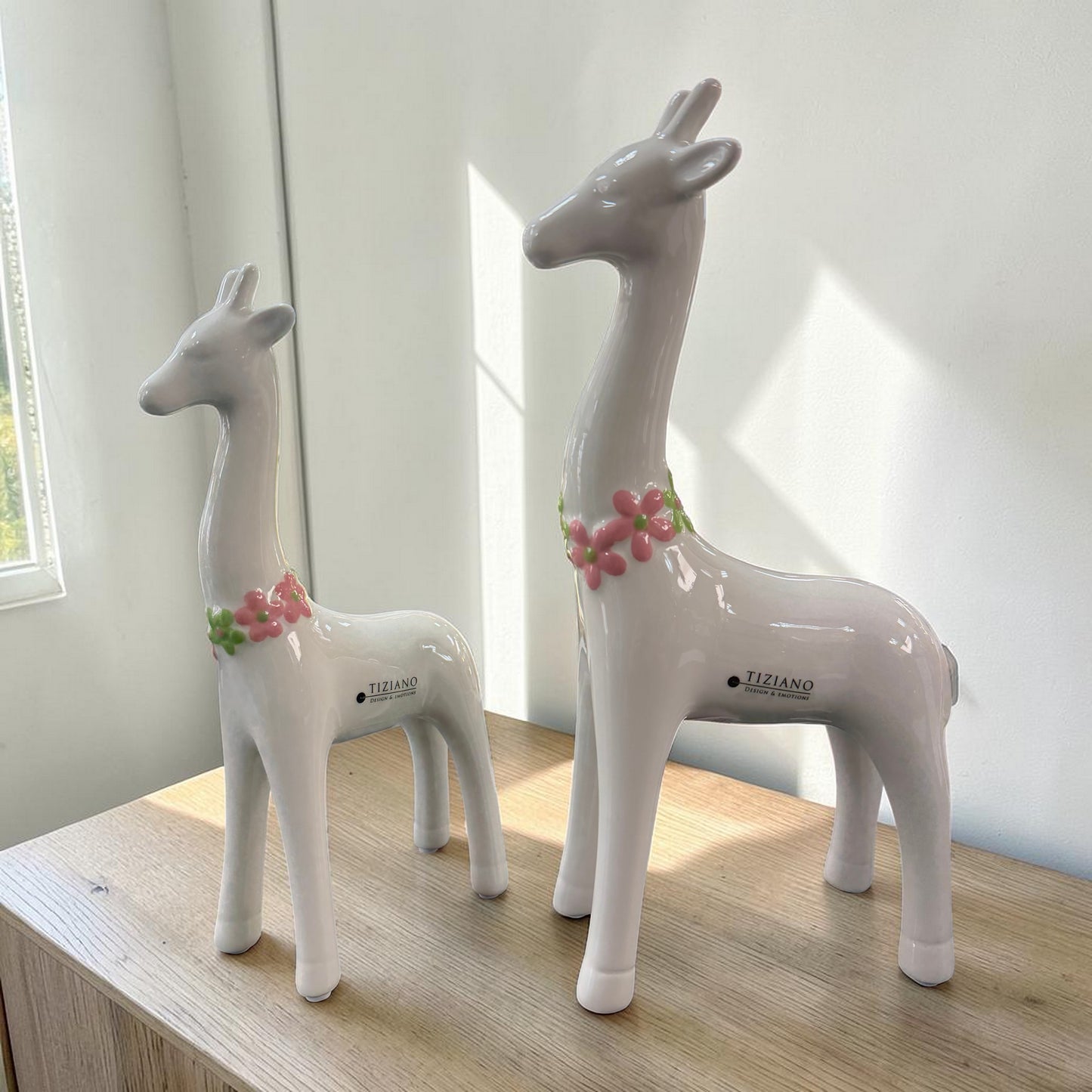 Zwei weiße Keramik-Giraffen mit langen Hälsen und rosa-grünem Blumenkranz stehen auf einer hellen Holzoberfläche im Sonnenlicht vor einer schlichten Wand.