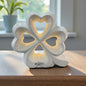 Großes weißes Keramik-Kleeblatt mit herzförmigen Aussparungen und warmer Beleuchtung auf einem Holztisch vor Fenster und Pflanze