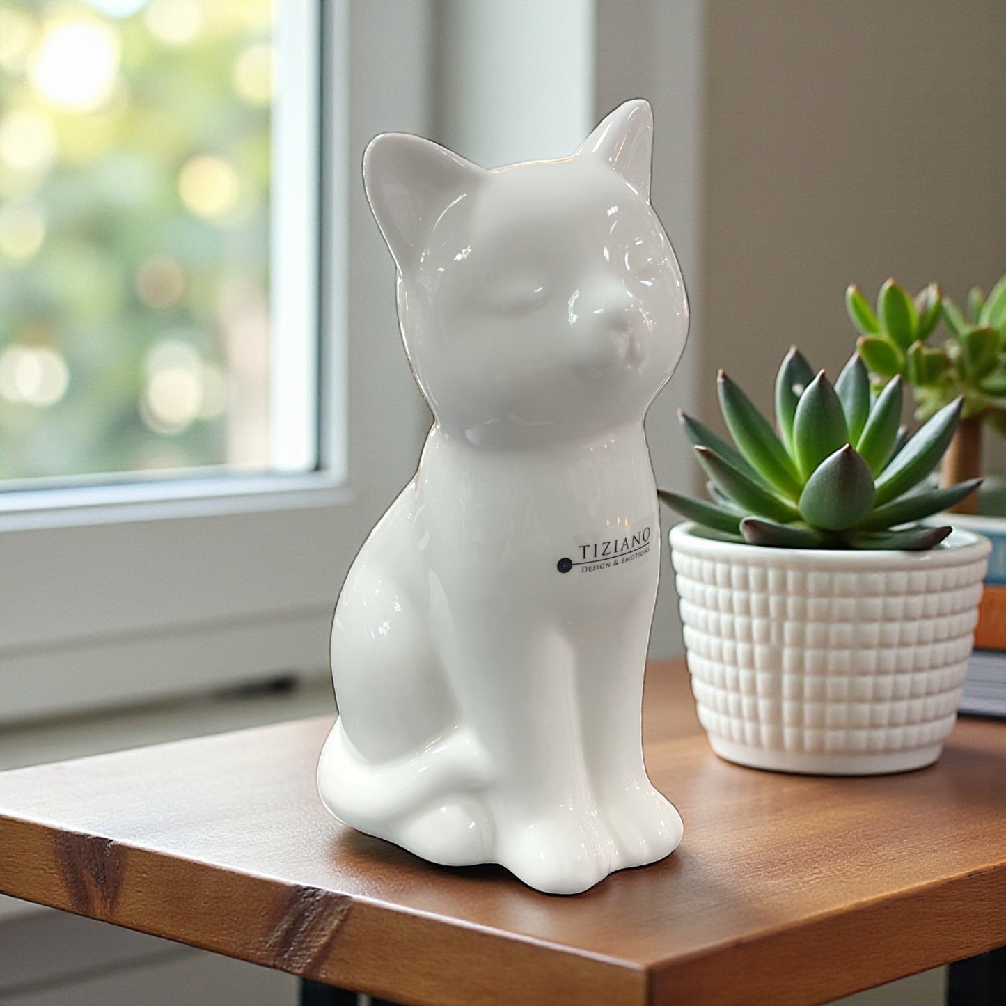 Eine weiße Keramik-Katzenfigur steht auf einem Holztisch vor einem Fenster, daneben ein kleiner Pflanztopf.