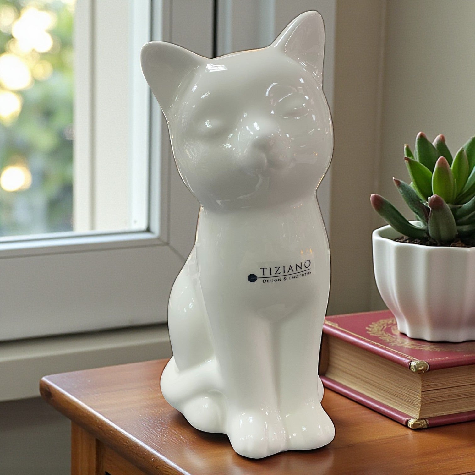 Eine große weiße Keramik-Katzenfigur steht auf einem Holztisch neben einem Buch und einer Topfpflanze vor einem Fenster.