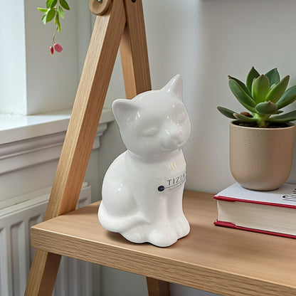 Eine kleine weiße Keramik-Katzenfigur steht dekorativ auf einem Holzregal neben einem Buch und einer Pflanze.