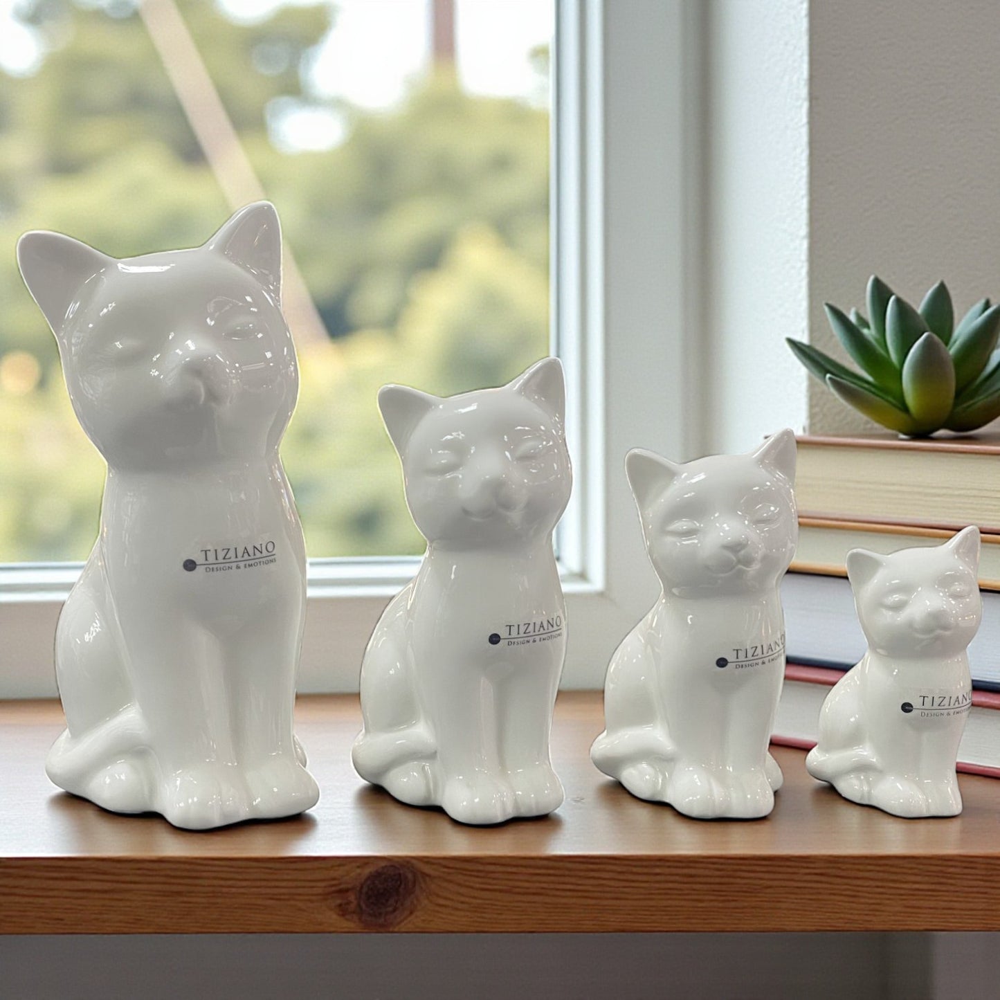 Vier weiße Keramik-Katzenfiguren in unterschiedlichen Größen stehen in einer Reihe auf einer Holzoberfläche vor einem Fenster.
