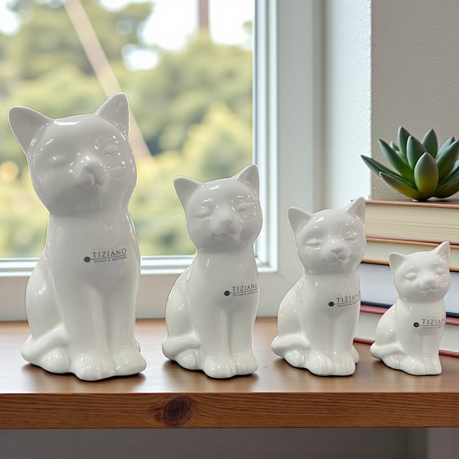 Vier weiße Keramik-Katzenfiguren in unterschiedlichen Größen stehen in einer Reihe auf einer Holzoberfläche vor einem Fenster.