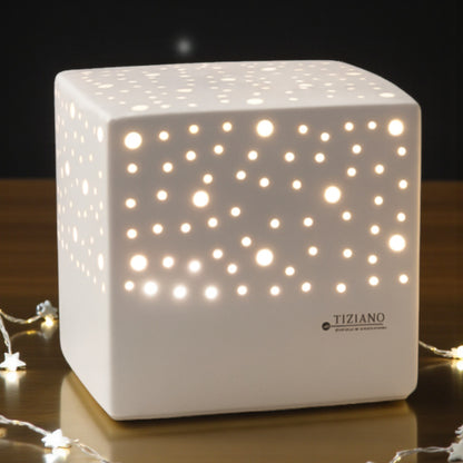 Quadratische, cremefarbene Keramiklampe mit Lochmuster steht auf einer Holzoberfläche. Durch die Öffnungen scheint warmes Licht, im Hintergrund ist eine dunkle Umgebung mit Lichterkette zu sehen.