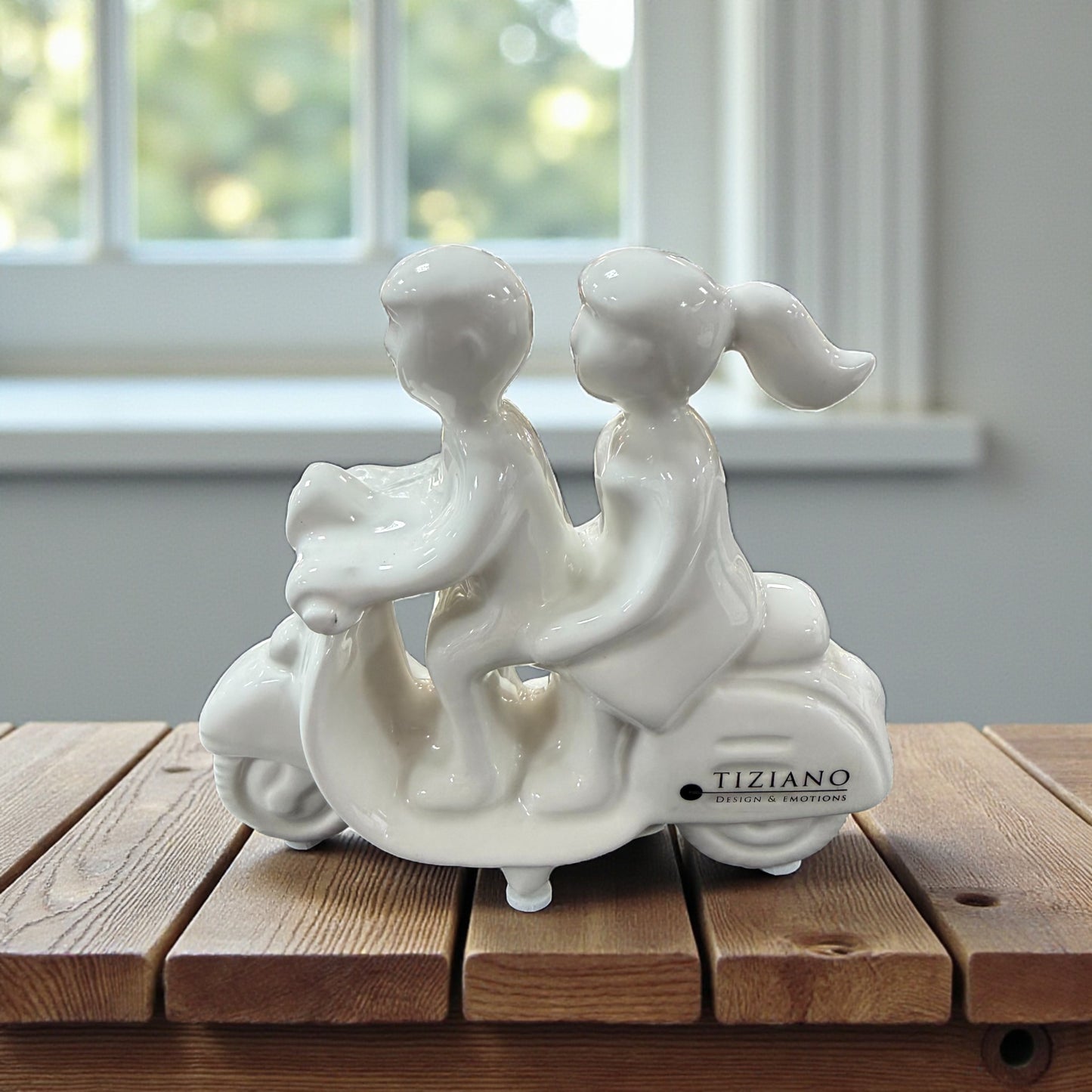 Weiße Keramikfigur eines Paares auf einem Roller auf einem Holztisch vor Fensterhintergrund.