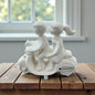 Weiße Keramikfigur eines Paares auf einem Roller auf einem Holztisch vor Fensterhintergrund.