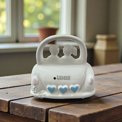 Weiße Keramikfigur eines Paares im Auto mit blauen Herzverzierungen auf einem Holztisch.