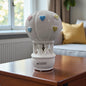 Weiße Keramikfigur mit Paar im Heißluftballon und bunten Herzen als Dekoration auf einem Tisch im Wohnraum.