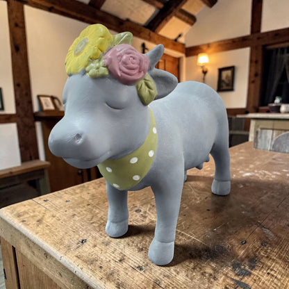 Eine graue Deko-Kuh aus Resin mit geschlossenen Augen, Blumenkranz auf dem Kopf und grün gepunktetem Halstuch steht auf einem Holztisch.