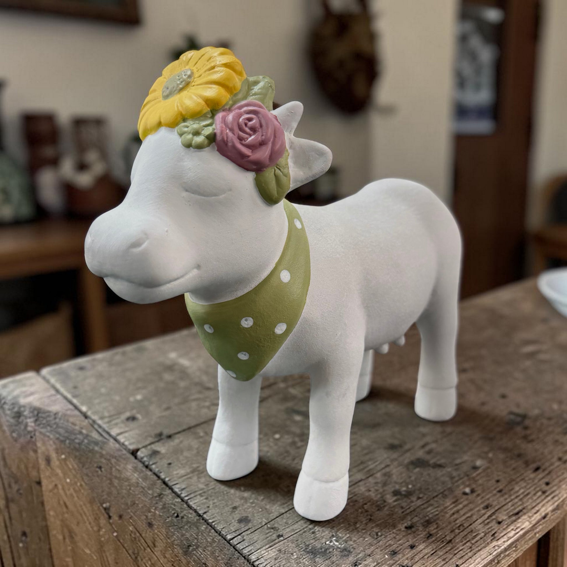 Eine weiße Deko-Kuh aus Resin mit gelber Blüte, rosa Rosen und grünem Halstuch steht auf einer rustikalen Holzoberfläche.