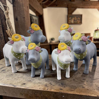Mehrere Deko-Kühe aus Resin in Weiß und Grau mit Blumenkranz und gepunktetem Halstuch stehen gemeinsam auf einem Holztisch.