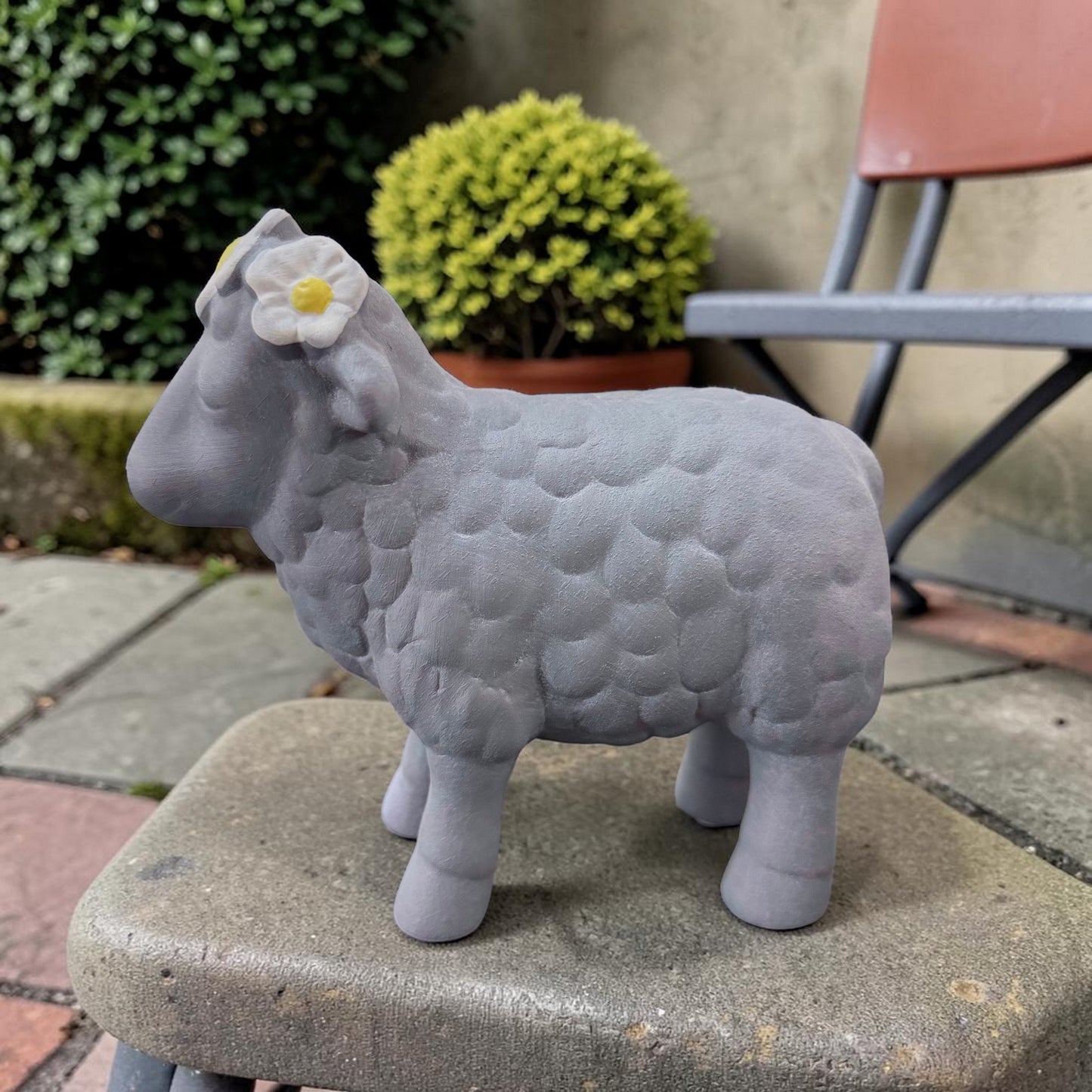 Ein graues Deko-Schaf aus Resin mit Blumenkranz steht auf einer Steinplatte vor einem Hintergrund mit Pflanzen und Gartenstuhl.