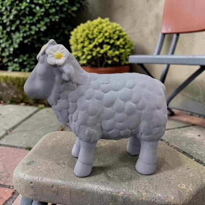Ein graues Deko-Schaf aus Resin mit Blumenkranz steht auf einer Steinplatte vor einem Hintergrund mit Pflanzen und Gartenstuhl.