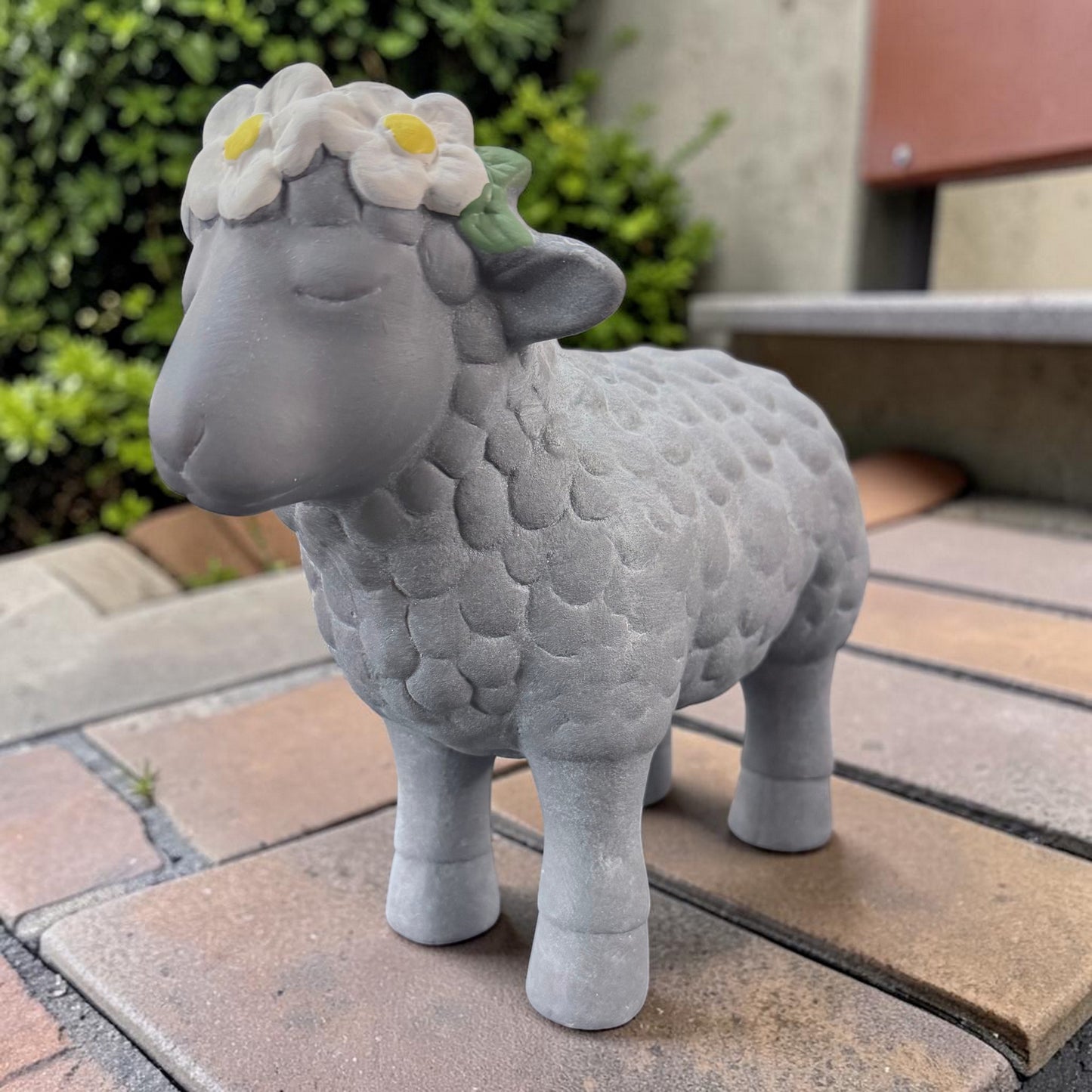 Ein graues Deko-Schaf aus Resin mit Blumenkranz steht auf einem gefliesten Untergrund vor einem unscharfen Gartenhintergrund.