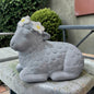 Ein sitzendes graues Deko-Schaf aus Resin mit weißen Blüten auf dem Kopf steht auf einer Steinplatte im Außenbereich.